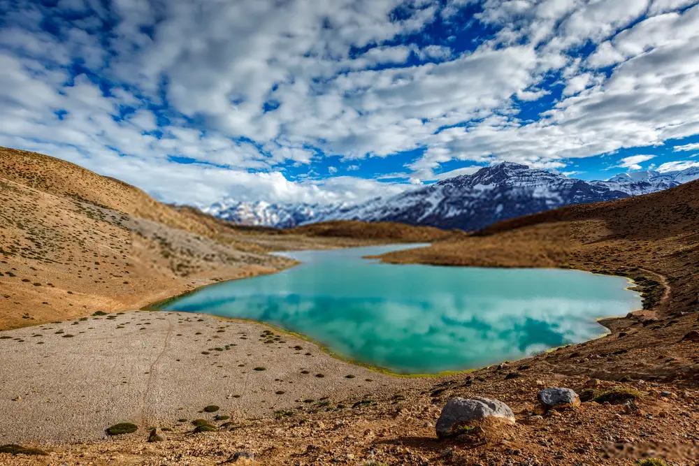 Panoramic view of the turquoise Dhankar Lake surrounded by the rugged, brown mountains of Spiti Valley under a clear blue sky.